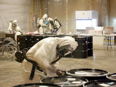 Workers in PPE examine large barrels for anthrax contamination.